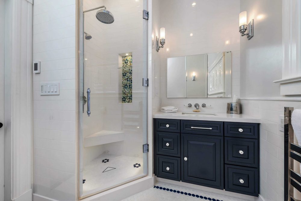 White tiled bathroom walk-in shower with stained glass niche and corner seat next to dark single sink vanity