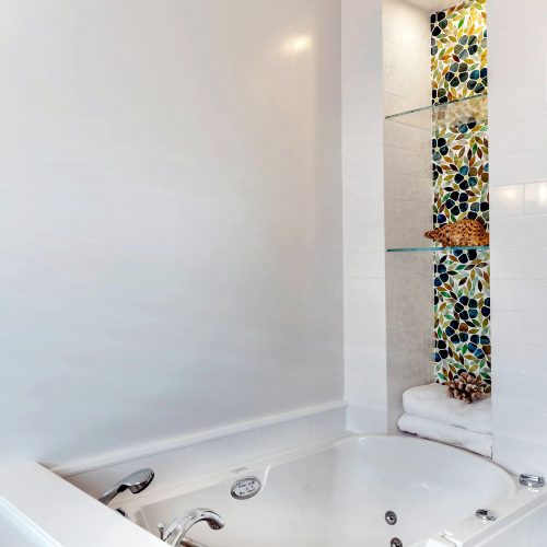 White tiled bathroom with sit tub and stained glass behind two glass shelves