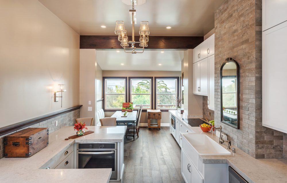 Long kitchen with white cabinets, stone backsplash, dark wood floors, 3 large windows at the end of the room near table