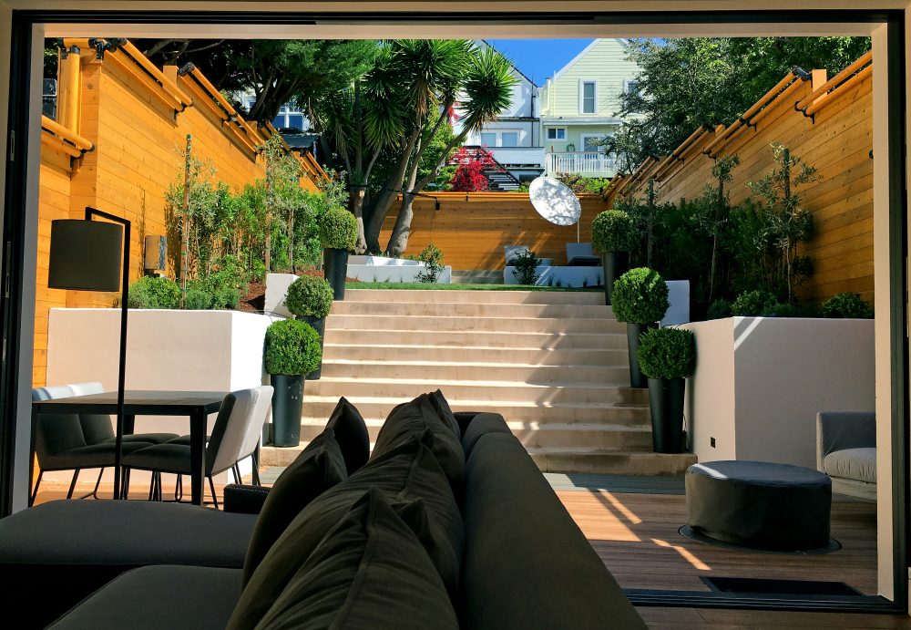 Looking out from a living room sofa to a long retangular backyard including patio with sitting and dining and 10 stone steps leading up to a grassy area surrounded by plants and trees