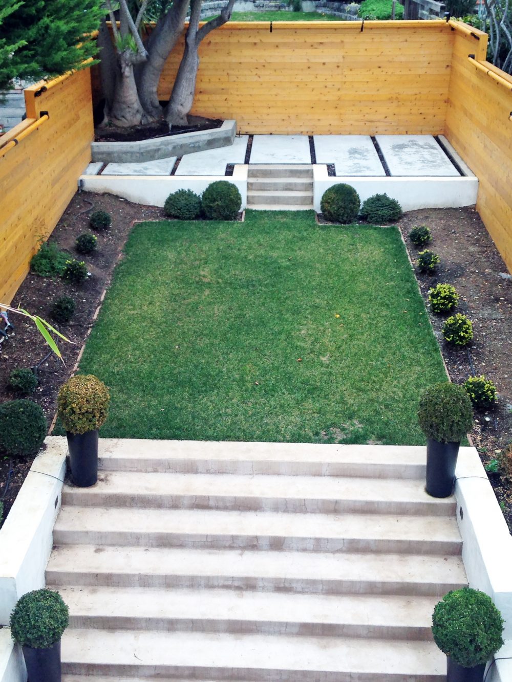 Arial view of long rectangular backyard and stone steps leading up to a grassy area and third elevated cement patio surrounded by slat fencing and landscaping