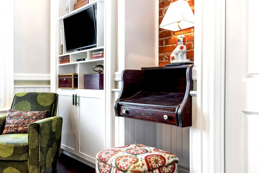 Built-in entertainment center next to an old style dark brown top portion of a secretary's desk with a lamp on top and a cushioned seat below