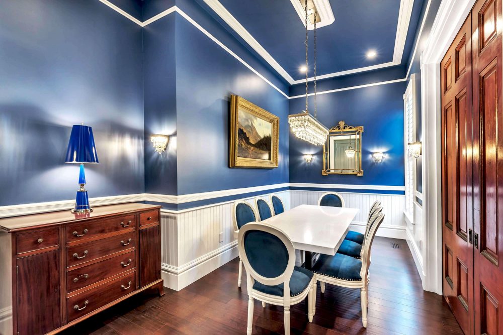 Fair Oaks dining room with blue walls and white crown molding; wood floor and door; wood server and white table with eight blue cushioned chairs